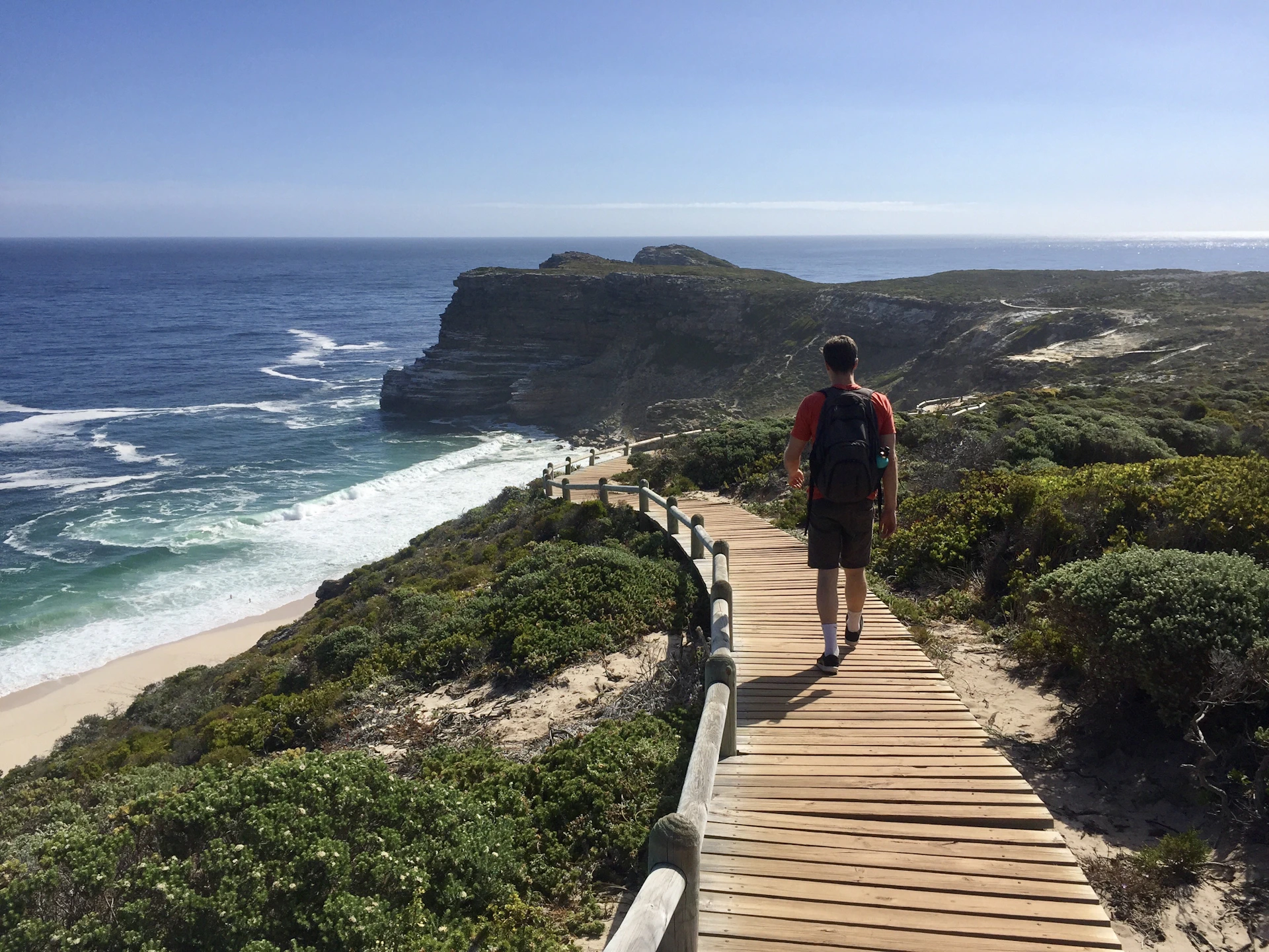 A sunny travel snapshot showing a backpacker walking along a scenic coastal trail with bright blue skies and calm ocean waves.
