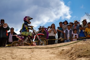 A motocross rider wearing protective gear and a helmet is on a dirt bike, participating in an event. A group of spectators, including adults and children, watch the action from behind a barrier. The scene is set outdoors under a partly cloudy sky.