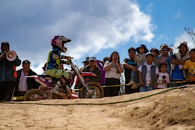 A motocross rider wearing protective gear and a helmet is on a dirt bike, participating in an event. A group of spectators, including adults and children, watch the action from behind a barrier. The scene is set outdoors under a partly cloudy sky.