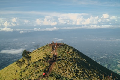 A group of hikers climbs a grassy hilltop path with expansive views of the landscape below. The sky is dotted with fluffy clouds, contributing to a serene and open atmosphere.