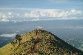 A group of hikers climbs a grassy hilltop path with expansive views of the landscape below. The sky is dotted with fluffy clouds, contributing to a serene and open atmosphere.