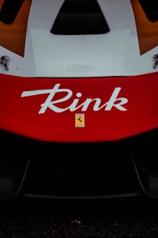 Close-up of a red and white racing car with the name 'Rink' prominently displayed in bold white letters. Below the name, a small emblem with the Ferrari logo is visible.