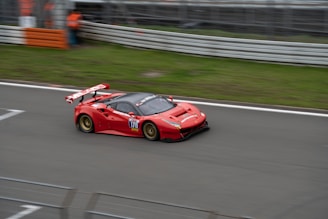 A red sports car with a large rear wing races along a track. The car has the number 178 and various sponsor stickers, and it appears to be in motion with some motion blur. The track is bordered by metal barriers and grass. There is a person in an orange outfit partially visible in the background near the barriers.