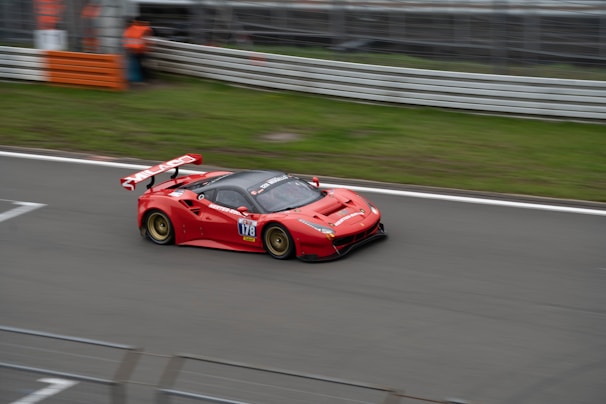 A red sports car with a large rear wing races along a track. The car has the number 178 and various sponsor stickers, and it appears to be in motion with some motion blur. The track is bordered by metal barriers and grass. There is a person in an orange outfit partially visible in the background near the barriers.