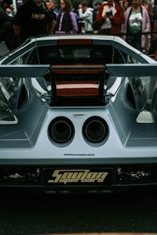 The rear view of a sleek and modern supercar with distinctive dual exhausts and a rear spoiler is surrounded by a crowd of people. The car appears to be part of a showcase or event, as indicated by the presence of a sign or license plate with 'Supercar Owners Circle'. The exterior has a glossy finish with aerodynamic contours.