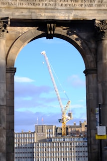 A large construction crane is seen framed by an archway, with a cityscape backdrop featuring a building labeled 'Leonardo Royal Hotel.' A blue sky with some clouds is visible in the background.