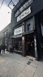 A storefront with the name 'Africa' displayed prominently on a sign above. The entrance has rustic wooden doors and there are two people walking on the sidewalk, with one person standing closer to the entrance. The shop window displays various goods, with greenery and plant motifs on the surrounding walls. The architecture is contemporary with a dark color scheme.