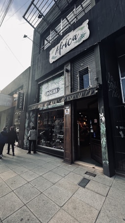 A storefront with the name 'Africa' displayed prominently on a sign above. The entrance has rustic wooden doors and there are two people walking on the sidewalk, with one person standing closer to the entrance. The shop window displays various goods, with greenery and plant motifs on the surrounding walls. The architecture is contemporary with a dark color scheme.