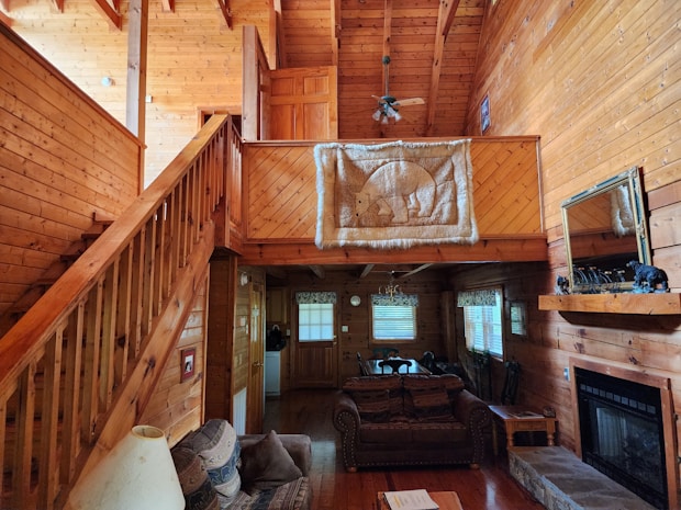 A cozy wooden interior featuring a loft area with a detailed bear tapestry hanging on the bannister. The space includes a staircase leading to the loft, a living room with a fireplace, a sofa, and a table with decorative items. The warm wood paneling covers the walls and ceiling, contributing to a rustic, cabin-like atmosphere.