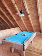 A billiard table with a bright blue felt cover is placed in a wooden room with a high angled ceiling. The table is equipped with a full set of billiard balls arranged in a triangle. Overhead, a ceiling fan with four lights hangs, complementing the rustic wooden decor.