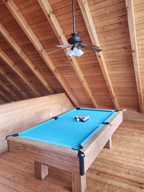 A billiard table with a bright blue felt cover is placed in a wooden room with a high angled ceiling. The table is equipped with a full set of billiard balls arranged in a triangle. Overhead, a ceiling fan with four lights hangs, complementing the rustic wooden decor.