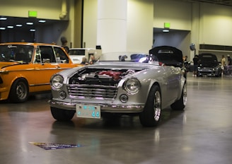 A classic silver convertible car with exposed engine components parked indoors, surrounded by other vintage vehicles. The background shows a spacious hall with people casually observing the cars on display. The lighting is artificial from overhead sources, giving a subtle shine to the vehicle's exterior.