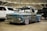 A weathered, vintage pickup truck with a rusted blue and brown exterior is prominently displayed in an indoor showroom. The truck has shiny chrome wheels and is parked among modern vehicles, including a white SUV and a black truck. A 'For Sale' sign is placed on the windshield, and the venue features industrial ceilings with visible beams and lighting.