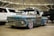 A weathered, vintage pickup truck with a rusted blue and brown exterior is prominently displayed in an indoor showroom. The truck has shiny chrome wheels and is parked among modern vehicles, including a white SUV and a black truck. A 'For Sale' sign is placed on the windshield, and the venue features industrial ceilings with visible beams and lighting.