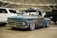 A weathered, vintage pickup truck with a rusted blue and brown exterior is prominently displayed in an indoor showroom. The truck has shiny chrome wheels and is parked among modern vehicles, including a white SUV and a black truck. A 'For Sale' sign is placed on the windshield, and the venue features industrial ceilings with visible beams and lighting.