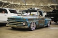 A weathered, vintage pickup truck with a rusted blue and brown exterior is prominently displayed in an indoor showroom. The truck has shiny chrome wheels and is parked among modern vehicles, including a white SUV and a black truck. A 'For Sale' sign is placed on the windshield, and the venue features industrial ceilings with visible beams and lighting.