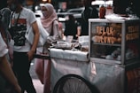 A street food cart with a sign reading 'Telor Gulung' is set up in a bustling outdoor area. The cart has eggs displayed in a glass case and cooking utensils are present. A group of people, some wearing face masks, are standing and walking nearby. One person is dressed in a pink hijab and white clothes, while another wears a white t-shirt with a graphic print.