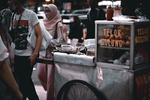A street food cart with a sign reading 'Telor Gulung' is set up in a bustling outdoor area. The cart has eggs displayed in a glass case and cooking utensils are present. A group of people, some wearing face masks, are standing and walking nearby. One person is dressed in a pink hijab and white clothes, while another wears a white t-shirt with a graphic print.