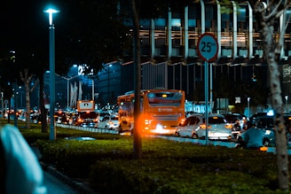A bustling urban street at night with heavy traffic. Cars and a bright orange bus are seen on the road, and a speed limit sign of 25 km/h is prominently visible. The street is well-lit with overhead lights, and there is lush greenery and trees along the sidewalk. The background comprises modern high-rise buildings, some with distinct architectural features.