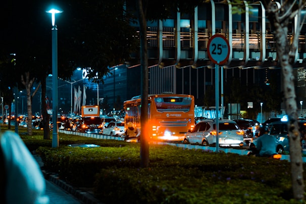 A bustling urban street at night with heavy traffic. Cars and a bright orange bus are seen on the road, and a speed limit sign of 25 km/h is prominently visible. The street is well-lit with overhead lights, and there is lush greenery and trees along the sidewalk. The background comprises modern high-rise buildings, some with distinct architectural features.