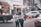 Two people are engaged with a mobile phone on a busy street, surrounded by parked vehicles. A black pickup truck is on the left with boxes loaded in the back. Several people are visible in the background, some engaged in various activities. Orange traffic cones are placed on the road, and buildings lined with signs and decorations form the backdrop.