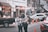 Two people are engaged with a mobile phone on a busy street, surrounded by parked vehicles. A black pickup truck is on the left with boxes loaded in the back. Several people are visible in the background, some engaged in various activities. Orange traffic cones are placed on the road, and buildings lined with signs and decorations form the backdrop.
