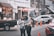 Two people are engaged with a mobile phone on a busy street, surrounded by parked vehicles. A black pickup truck is on the left with boxes loaded in the back. Several people are visible in the background, some engaged in various activities. Orange traffic cones are placed on the road, and buildings lined with signs and decorations form the backdrop.
