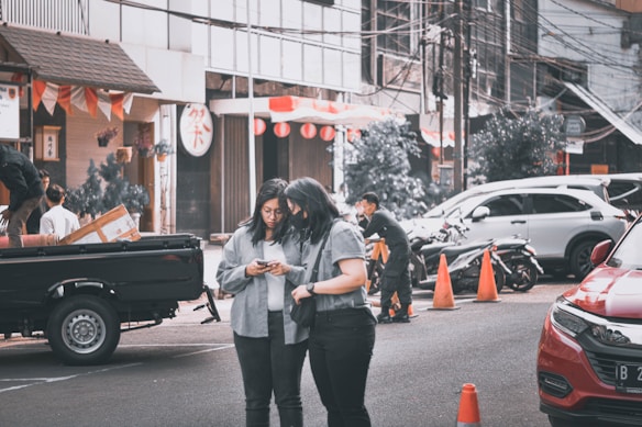 Two people are engaged with a mobile phone on a busy street, surrounded by parked vehicles. A black pickup truck is on the left with boxes loaded in the back. Several people are visible in the background, some engaged in various activities. Orange traffic cones are placed on the road, and buildings lined with signs and decorations form the backdrop.