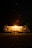 A well-lit camping trailer is positioned in a dark, wooded area at night. Warm light radiates from the trailer's window, creating a cozy atmosphere. It sits on a patch of grass surrounded by tall trees, with a faint glow in the sky hinting at distant stars.