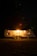 A well-lit camping trailer is positioned in a dark, wooded area at night. Warm light radiates from the trailer's window, creating a cozy atmosphere. It sits on a patch of grass surrounded by tall trees, with a faint glow in the sky hinting at distant stars.