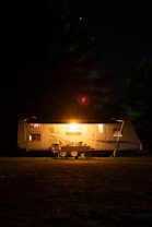A well-lit camping trailer is positioned in a dark, wooded area at night. Warm light radiates from the trailer's window, creating a cozy atmosphere. It sits on a patch of grass surrounded by tall trees, with a faint glow in the sky hinting at distant stars.