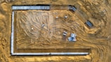 An aerial view of a construction site with visible tracks from construction vehicles on the sandy ground. Concrete slabs and construction materials are strategically placed around the site. The area is clear and defined by the surroundings of the site with materials spread out.