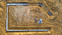 An aerial view of a construction site with visible tracks from construction vehicles on the sandy ground. Concrete slabs and construction materials are strategically placed around the site. The area is clear and defined by the surroundings of the site with materials spread out.