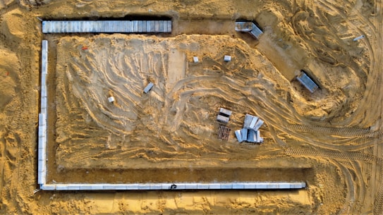 An aerial view of a construction site with visible tracks from construction vehicles on the sandy ground. Concrete slabs and construction materials are strategically placed around the site. The area is clear and defined by the surroundings of the site with materials spread out.