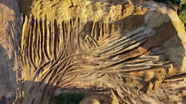 Aerial view of a construction or mining site with numerous parallel and intersecting tire tracks forming a pattern on the dusty surface. The ground is predominantly brown with shadowed areas, showing signs of heavy vehicle activity. Sparse greenery appears on the edges, contrasting with the earth tones.