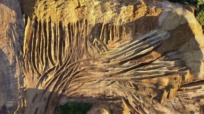 Aerial view of a construction or mining site with numerous parallel and intersecting tire tracks forming a pattern on the dusty surface. The ground is predominantly brown with shadowed areas, showing signs of heavy vehicle activity. Sparse greenery appears on the edges, contrasting with the earth tones.