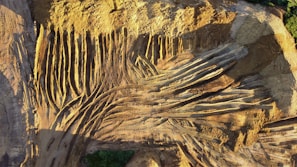Aerial view of a construction or mining site with numerous parallel and intersecting tire tracks forming a pattern on the dusty surface. The ground is predominantly brown with shadowed areas, showing signs of heavy vehicle activity. Sparse greenery appears on the edges, contrasting with the earth tones.