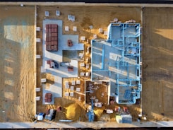 An aerial view of a construction site with a partially built structure on a sandy ground. Blue steel frames outline the layout of the building while red and white materials are neatly stacked around the premises. Shadows cast at an angle suggest early morning or late afternoon light.