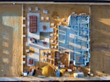 An aerial view of a construction site with a partially built structure on a sandy ground. Blue steel frames outline the layout of the building while red and white materials are neatly stacked around the premises. Shadows cast at an angle suggest early morning or late afternoon light.