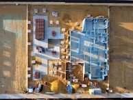 An aerial view of a construction site with a partially built structure on a sandy ground. Blue steel frames outline the layout of the building while red and white materials are neatly stacked around the premises. Shadows cast at an angle suggest early morning or late afternoon light.
