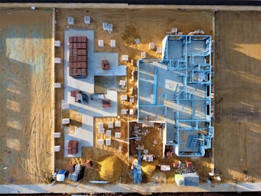 An aerial view of a construction site with a partially built structure on a sandy ground. Blue steel frames outline the layout of the building while red and white materials are neatly stacked around the premises. Shadows cast at an angle suggest early morning or late afternoon light.