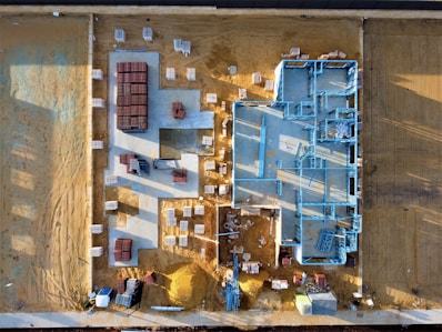 An aerial view of a construction site with a partially built structure on a sandy ground. Blue steel frames outline the layout of the building while red and white materials are neatly stacked around the premises. Shadows cast at an angle suggest early morning or late afternoon light.