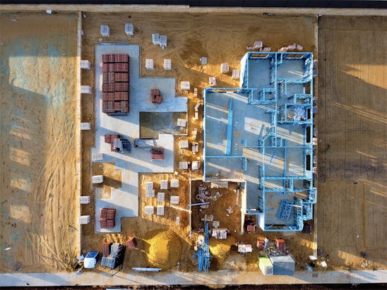 An aerial view of a construction site with a partially built structure on a sandy ground. Blue steel frames outline the layout of the building while red and white materials are neatly stacked around the premises. Shadows cast at an angle suggest early morning or late afternoon light.