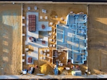 An aerial view of a construction site with a partially built structure on a sandy ground. Blue steel frames outline the layout of the building while red and white materials are neatly stacked around the premises. Shadows cast at an angle suggest early morning or late afternoon light.