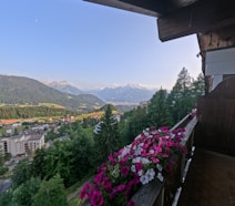 A picturesque view from a balcony overlooking a valley containing a small town, surrounded by lush green trees and distant mountains under a clear sky. Pink and white flowers decorate the balcony railing.