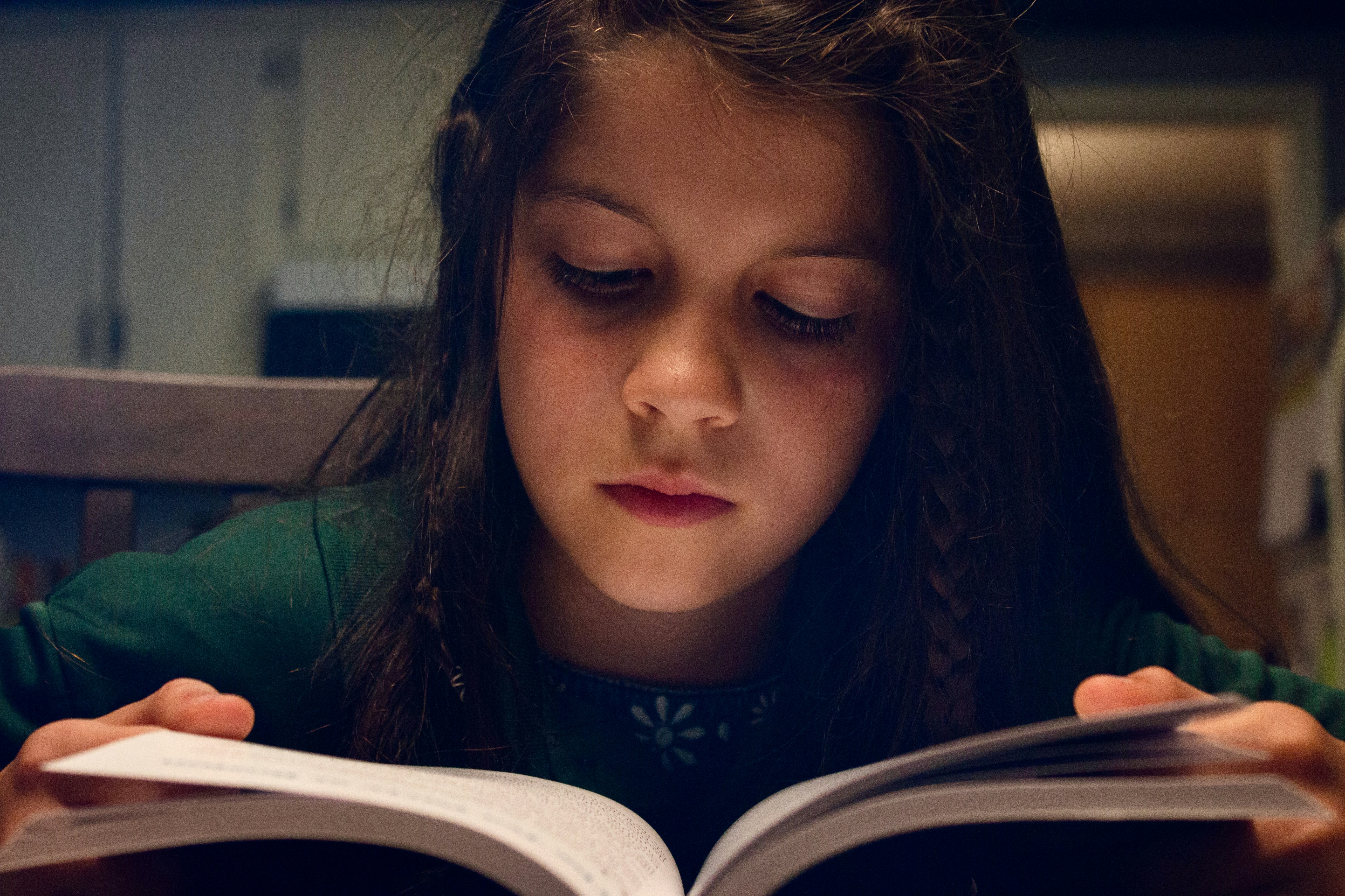 Young girl reading a book in a dimly lit room, focused expression, warm amber light