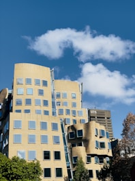 A modern, uniquely designed building with numerous windows under a clear blue sky with scattered white clouds. The architecture features irregular shapes and curves. In the background, a tall building labeled 'UTS' is visible. Green trees are present in the foreground.