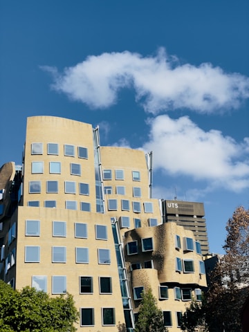A modern, uniquely designed building with numerous windows under a clear blue sky with scattered white clouds. The architecture features irregular shapes and curves. In the background, a tall building labeled 'UTS' is visible. Green trees are present in the foreground.
