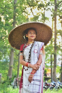 A woman stands in a forested area wearing traditional attire, including a large woven hat and a saree. The sunlight filters through the tall green trees, creating a serene and natural atmosphere. She is adorned with jewelry and colorful bangles, and her expression is peaceful and content.