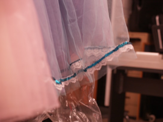 A close-up view of fabric with layers of pastel blue and pink tulle. The edges are adorned with lace and a blue sequin trim. Part of a garment is visible against a blurred background, suggesting a studio or dressing room setting.